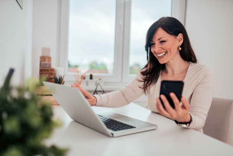 Mulher sorridente com celular em uma mão e notebook em cima da mesa de escritório, satisfeita com a facilidade de emitir aviso de férias.